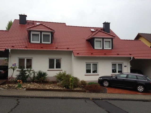 Ein weißes Doppelhaus mit einem leuchtend rot beschichteten Ziegeldach, zwei großen Dachgauben und zwei dunklen Schornsteinen unter einem bewölkten Himmel.