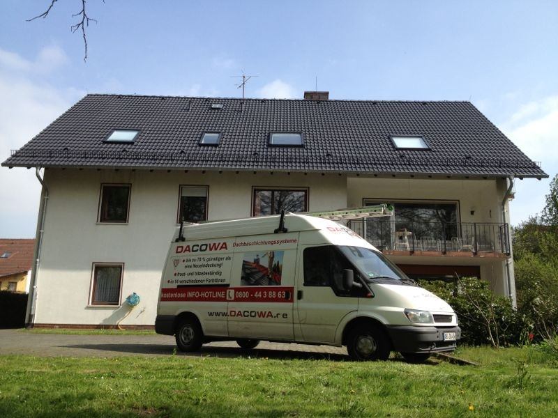 Ein weißer Transporter der Firma „DACOWA Dachbeschichtungssysteme“ parkt auf einer Rasenfläche vor einem zweistöckigen Wohnhaus mit großem, anthrazitfarbenem Satteldach und mehreren Dachfenstern.