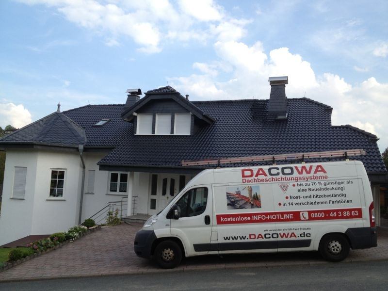 Ein weißer Firmenwagen der Firma „DACOWA Dachbeschichtungssysteme“ steht in einer gepflasterten Einfahrt vor einem großen, zweistöckigen Einfamilienhaus mit frisch beschichtetem, dunkelblauem Ziegeldach.
