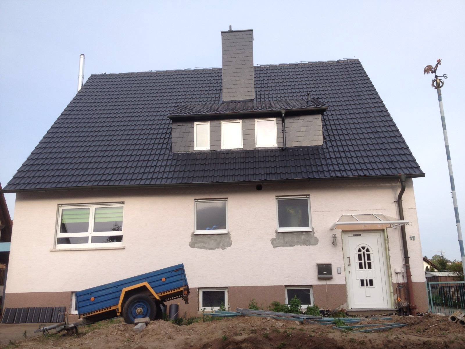 Modernisiertes Wohnhaus mit einer glänzend schwarzen Dachbeschichtung und einer farblich passenden Schiefergaube; im Vordergrund stehen ein blauer Arbeitsanhänger und Baumaterialien vor der hellen Fassade.
