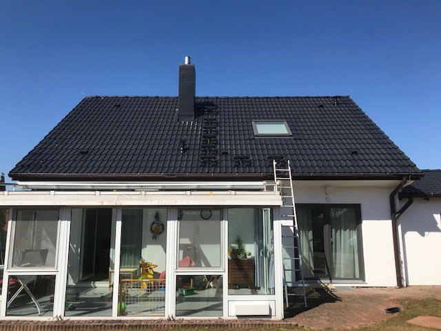 Modernes weißes Einfamilienhaus mit Wintergarten während einer Dachbeschichtung; das tiefschwarze Satteldach glänzt frisch versiegelt unter blauem Himmel, eine Anliegeleiter lehnt am Dachrand.
