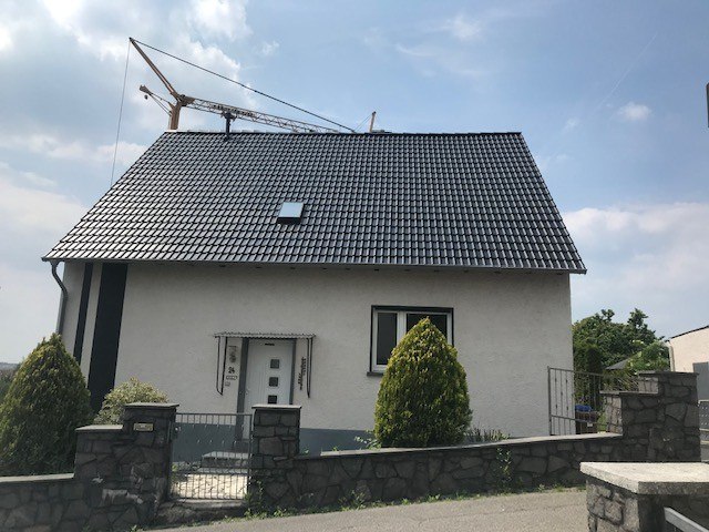 Einfamilienhaus mit einer glänzenden, anthrazitfarbenen Dachbeschichtung auf einem Steildach, fotografiert bei hellem Tageslicht mit einem Baukran im Hintergrund.