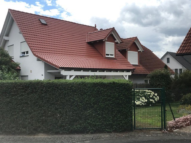 Gepflegtes Einfamilienhaus mit einem farbintensiven, tiefroten Satteldach und zwei markanten Giebelgauben, umgeben von einer hohen Hecke und einem Garten.