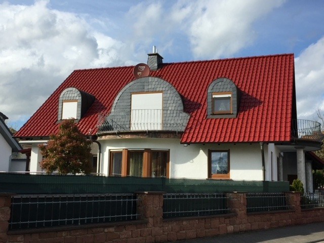 Modernisiertes Einfamilienhaus mit einer farbintensiven Dachbeschichtung in Rubinrot, kombiniert mit grauen Schiefergauben und einem markanten Rundbogen-Balkon.