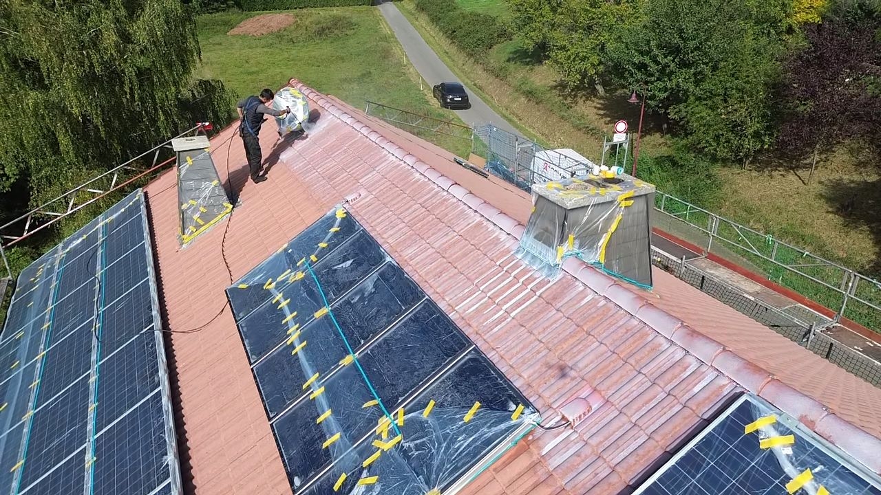 Luftaufnahme einer Dachbeschichtung: Ein Handwerker trägt mit einem Airless-Gerät rote Farbe auf ein Ziegeldach auf. Photovoltaikanlagen und Kamine sind zum Schutz sorgfältig mit Folie abgeklebt.